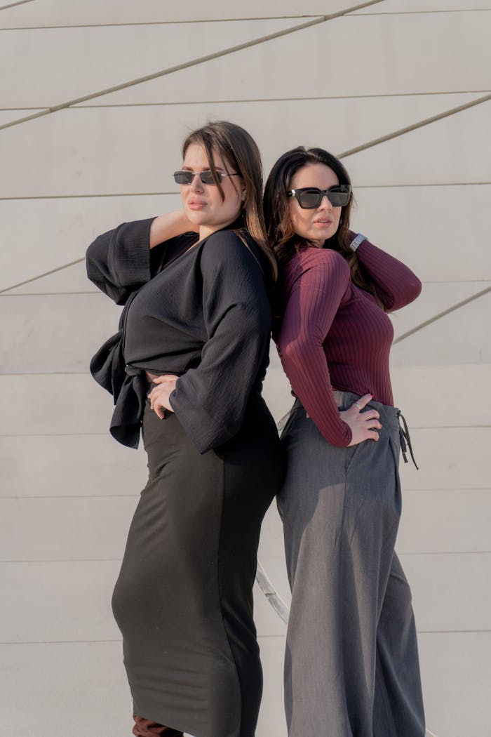 Two stylish women with sunglasses pose confidently against a modern wall outdoors.